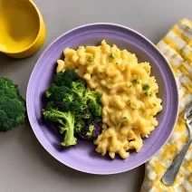 Fotografia em tons de roxo e amarelo com um prato roxo ao centro. Dentro do prato existe uma receita de Mac'n Cheese com brócolis.