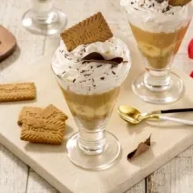 Fotografia em tons de marrom claro com duas taças de vidro ao centro. Dentro de cada taça existe uma receita de banoffee com camadas de biscoito TOSTINES, banana, doce de leite e chantilly.