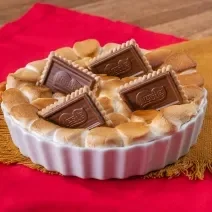 Fotografia de uma forma branca de vidro com s'mores que foi ao forno, e por cima, quatro biscoitos de chocobiscuit. A forma está apoiada sobre um paninho de tom marrom.