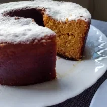 Fotografia em tons de bege em uma bancada com toalha cinza, um prato branco redondo raso, um bolo de milho cortado ao meio com açúcar polvilhado em cima.