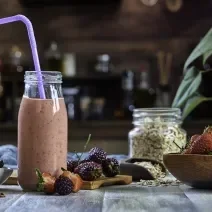 Fotografia de uma bebida densa de morango com leite MOÇA e neston que está em uma garrafa de vidro decorativa, com um canudo roxo e frutas no fundo.