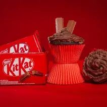 Fotografia em tons de vermelho em uma bancada de madeira de cor vermelha. Ao centro, um cupcake em pé em cima de uma forminha. AO lado direito, um cupcake deitado e ao lado esquerdo, 2 embalagens de kitkat ao laite.