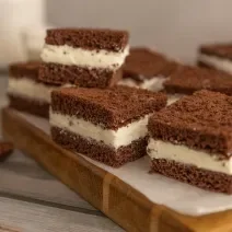 Fotografia de pedaços quadrados de bolo de chocolate recheado com creme de limão, e estão sobre um papel manteiga, apoiado em uma tábua de madeira.