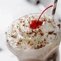 Fotografia de um copo de vidro longo com uma bebida gelada e cremosa de sorvete de chocolate, leite e chantilly. Em cima da bebida, tem raspas de chocolate e um canudo de cores alternadas e uma cereja.