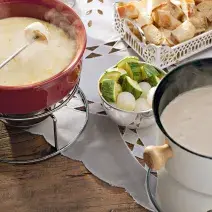 recipientes nas cores vermelho e branco ambos comporta o fondue de queijo, outros recipientes estão com os acompanhamentos para o fondue como pão, presunto entre outros.