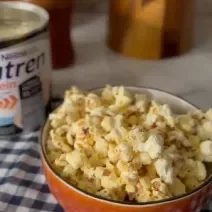 Foto da receita de pipoca doce proteica servida em um bowl laranja sobre uma mesa de mármore branca com um paninho azul xadrez. Ao fundo há uma embalagem de nutren protein e utensílios de madeira.