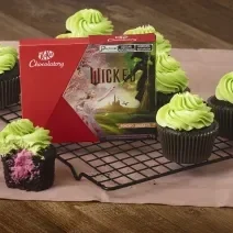 Foto da receita de Cupcake Wicked, servidos em uma grade. Eles são bolinhos escuros, recheados com um creme rosa e com uma cobertura verde clara. Em meio a eles está uma caixa de KitKat Wicked.