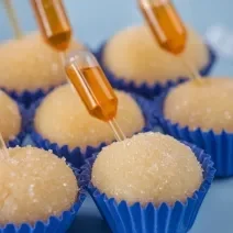 Fotografia de sete forminhas azuis juntas com brigadeiro de pudim, em cada unidade tem uma mini pipeta espetada com calda de caramelo. Os brigadeiros estão apoiados em um apoio decorativo azul claro.