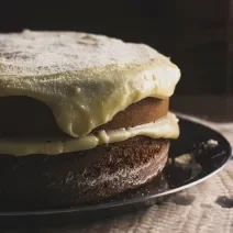 Fotografia de um bolo de abóbora com creme de coco por cima. O bolo está recheado com o mesmo creme, e está sobre um prato raso preto.