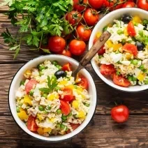 Fotografia vista de cima de dois recipientes brancos fundos com arroz e pimentões preparado no micro-ondas com MAGGI Fondor e Creme de Leite NESTLÉ. Em um dos recipientes têm um talher apoiado. Ao redor tem tomates cereja sobre uma mesa de madeira.