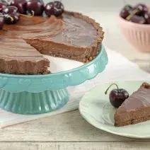 Fotografia em tons de marrom, branco, azul e rosa de uma bancada cinza vista de frente. Um recipiente azul contém uma torta de chocolate com varias cerejas e com uma fatia retirada, ao lado um prato branco com a fatia de torta e uma cereja do lado.