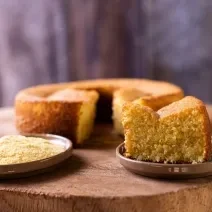 Foto da receita de bolo de milho de liquidificador, redondo, dourado, com uma fatia mais à frente em um prato e ao lado um punhado de fubá em outro prato. Ao fundo é possível ver o bolo inteiro