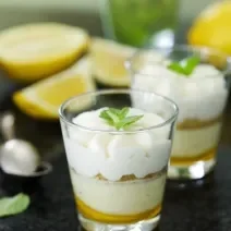 Fotografia de dois copinhos de vidro com farofa de biscoito e creme de limão sobre uma mesa de mármore preta. Ao fundo, duas metades de um limão siciliano, muitas folhas de hortelã e uma colher de sobremesa.