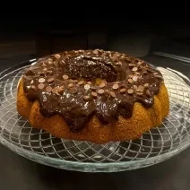 Fotografia de um bolo de cenoura sem glúten sobre um apoio de vidro decorativo. Por cima do bolo com furo no centro tem uma cobertura de chocolate com gotas de chocolate.