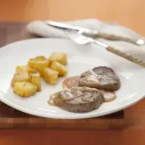 Fotografia em tons de branco e marrom de uma mesa de madeira, vista de lado, ao centro um prato redondo branco, com o filé regado com molho e batatas assadas para acompanhar. Ao lado talheres e um guardanapo de linho em tons beges.