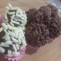 Fotografia de oito brigadeiros dentro de forminhas rosa. Seis brigadeiros foram enrolados em confeitos de chocolate preto, e dois deles foram enrolados em confeitos brancos. As forminhas estão sobre uma forma de madeira.