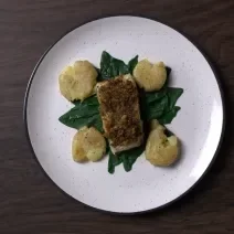 Fotografia em tons de branco com um prato branco ao centro. Dentro do prato existe uma receita de bacalhau acompanhado de batatas amassadas e espinafre.