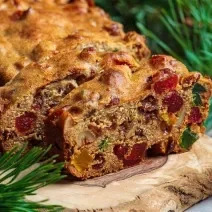 Fotografia de um bolo de frutas cristalizadas e secas com whisky e especiarias, feito na forma pequena de bolo inglês. Tem duas fatias cortadas na frente do restante do bolo, que está sobre um apoio de madeira, ao lado de paus de canela.