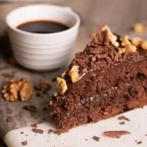 Fotografia de uma fatia de bolo feito com Chocolate NESTLÉ CLASSIC Meio Amargo, nozes e Cobertura de Chocolate ao Leite GAROTO, com raspas de chocolate e castanhas por cima. Ao lado da fatia tem uma xícara pequena branca com café.