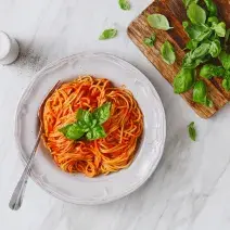 fotografia em tons de branco e vermelho de uma bancada branca vista de cima. Ao centro um prato branco com macarrão espaguete e molho de tomate, e ao lado uma tábua com folhas.