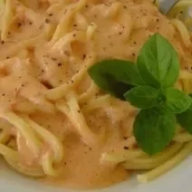 Fotografia em tons de vermelho, em uma bancada de madeira de cor marrom. Ao centro, um prato branco contendo o macarrão.