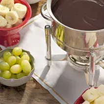 Fotografia em tons de verde e vermelho em uma bancada de madeira, uma toalhinha cinza, potinhos vermelhos com banana, uva, morango e abacaxi dentro deles. Ao centro, uma panela com a fondue de chocolate amargo dentro.