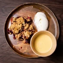 Fotografia vista de cima de um prato raso de vidro marrom, com um crumble de de frutas com creme inglês em um recipiente e chantilly de iogurte ao lado. O prato está sobre uma mesa de madeira.