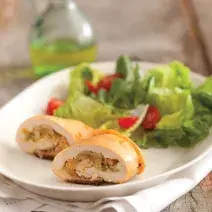 Fotografia em tons de verde em uma bancada de madeira com um prato oval branco com o enroladinho de frango com salada de alface e tomate cereja.