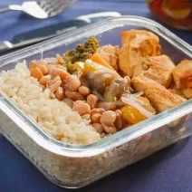 Marmita em pote de vidro transparente, com arroz integral, feijão, cubos de frango dourados e legumes assados coloridos (brócolis, cenoura, pimentões). Tudo bem distribuído, destacando cores vibrantes e textura fresca.