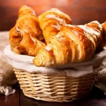 Fotografia de cestinhas de palha com um pano de tom bege. Dentro da cestinha no centro da foto, tem vários pães tipo croissant.