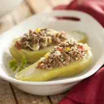 Fotografia em tons de branco, verde e vinho, ao centro refratário branco com duas metades de chuchu com carne moída, ao lado guardanapo vinho, pote branco ao fundo, tudo sobre bancada de madeira envelhecida.