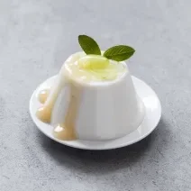 Fotografia de uma mousse de uva verde, com um creme e uma unidade de uva em cima, e para decorar, duas folhas de hortelã. A sobremesa está em um prato pequeno de sobremesa na cor branco, que está sobre uma mesa cinza.