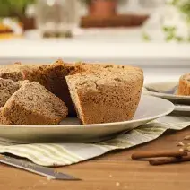 Fotografia em tons de marrom em uma mesa de madeira com um prato raso branco apoiado em um pano cinza listrado em bege e o bolo de maçã e nozes. Ao lado, um prato com um pedaço do bolo e paus de canela espalhadas pela mesa.