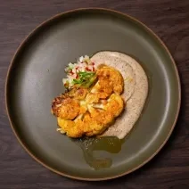 Fotografia em tons de marrom com um prato cinza ao centro. Em cima do prato existe uma receita de couve-flor tostada na panela com creme de castanhas e vinagrete