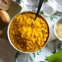 Fotografia vista de cima de um refratário fundo branco com arroz cremoso com açafrão. Ao lado direito, dois pratos e dois copos brancos, um bowl pequeno branco com queijo ralado, e no outro lado, um prato quadrado verde com alguns pães.