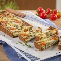 Fotografia em tons branco, dourado e vermelho, a direita e ao alto tomates-cereja e folhas de tomilho, ao centro travessa branca com quiche assada e fatiada, sobre papel manteiga e guardanapo azul em bancada de madeira.