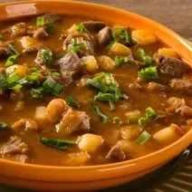 Foto aproximada de um prato marrom com a receita de carne de panela com mandioca dentro, em um caldo grosso e marrom, com pedaços de carne, mandioca e cheiro-verde picado