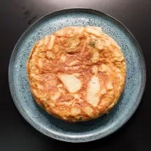 Fotografia em tons de preto com um prato azul ao centro. Dentro do prato existe uma receita de Tortilha Espanhola, uma receita feita com ovos e batatas cortadas de forma fina.