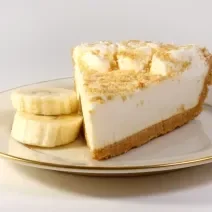 Fotografia de uma fatia de torta de banana e leite MOÇA, com uma farofinha de biscoito e rodelas de banana por cima. A torta está em um prato de vidro branco com as bordas na cor dourado, com duas rodelas de banana ao lado da torta, sobre uma mesa branca.
