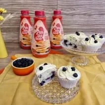 Fotografia em tons de amarelo com dois recipientes de vidro ao centro. Em cima dos recipientes existe vários cupcakes recheados com blueberry e coberto com um creme branco feito de GALAK.