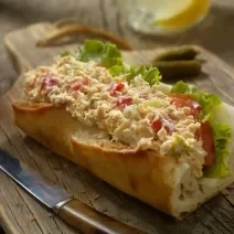 Fotografia de um sanduíche no pão francês com tomate, patê, queijo e alface, sobre uma tábua de madeira escura. Ao lado, uma faca com o cabo marrom.
