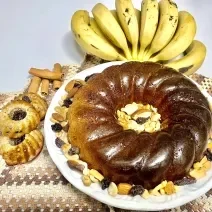 Fotografia de um prato branco raso e grande com o bolo inteiro. Dentro do espaço do bolo e em volta tem castanhas e frutas secas decorando.