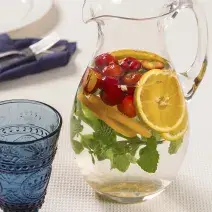 fotografia em tons de branco e azul de uma bancada vista de cima, uma jarra transparente com agua, pedaços de laranja, acerola e hortelã e ao lado um copo cor azul para servir