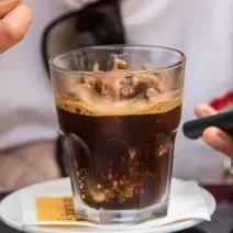 Fotografia de uma granita feita de café, leite e gelo que está em um copo de vidro, o qual está apoiado sobre um miniprato de vidro branco. Ao fundo, tem alguém segurando uma colher com a sobremesa.