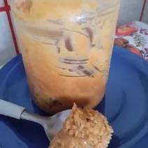 Fotografia de um pote de vidro com doce de leite caseiro com coco sobre um prato raso azul. Na frente do vidro tem uma colher com doce de leite e coco ralado.