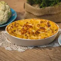 Fotografia em tons de amarelo em uma bancada de madeira clara, um paninho de renda e um recipiente branco oval com a couve-flor gratinada feita na AirFryer Mondial dentro dele. Ao lado, um prato com couve-flor crua, um outro prato com uma fatia.