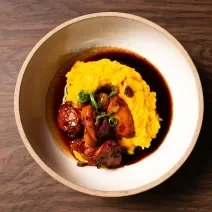 Fotografia vista de cima de uma receita com arroz cremoso na cor amarela sobre de um molho com shoyu. Por cima do arroz tem uns pedaços de frango caramelizados, e cebolinha para enfeitar.