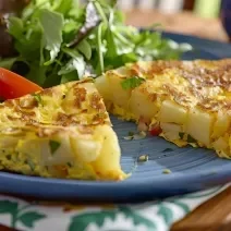 Foto aproximada da receita de omelete com batata, servida em um prato e dividia ao meio, podendo ver as batatas no centro. No prato há uma salada verde e algumas fatias de tomate