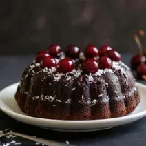 Fotografia de um bolo de nescau, feito na forma redonda com furo no meio, com cereja na massa e várias cerejas por cima do bolo, que está sobre um prato fundo branco. O prato está sobre uma mesa preta, que tem várias cerejas juntas.