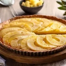 imagem de uma torta redonda com rodelas de abacaxi assadas por cima. A torta permanece em uma forma de tortas com as laterais chanfradas e está sobre uma tábua de mandeira. ao fundo, um pedaço da torta fica sobre um prato branco e ao lado um abacaxi.